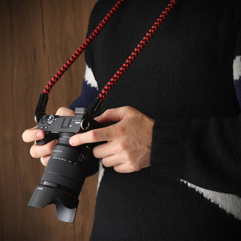 May include: A black camera with a red and black camera strap. The camera has a lens with a black barrel and a silver ring around the lens.