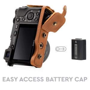 Pu&ograve; includere: Una custodia per fotocamera in pelle marrone con una patta che si apre per rivelare una fotocamera nera con un obiettivo. La custodia ha una tracolla e uno scomparto per batterie con una batteria etichettata "InfoLITHIUM W".