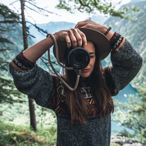 May include: A person wearing a grey sweater with a colorful pattern and a brown hat is holding a black camera with a brown strap. The person is standing in front of a green forest and a body of water.