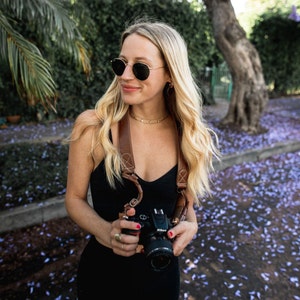 May include: A woman wearing sunglasses and a black tank top holds a black camera with a brown leather strap. The camera is in focus, and the woman is slightly out of focus. The background is a blurry image of a tree and purple flowers.