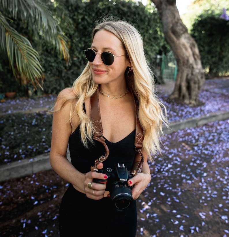 May include: A woman wearing sunglasses and a black tank top holds a black camera with a brown leather strap. The camera is in focus, and the woman is slightly out of focus. The background is a blurry image of a tree and purple flowers.