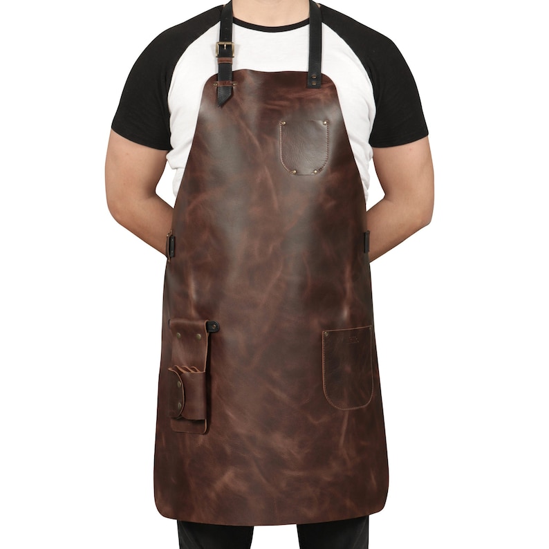 May include: A brown leather apron with adjustable straps, two pockets, and a small tool loop. The apron is being worn by a person, but the person is not visible in the image.