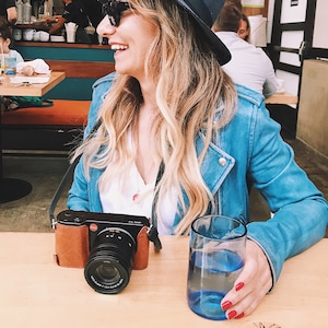 Op de afbeelding: Een vrouw met een blauwe denimjas en een zwarte hoed houdt een bruine leren cameratas vast met een zwarte camera lens. Ze zit aan een tafel met een glas blauwe vloeistof.