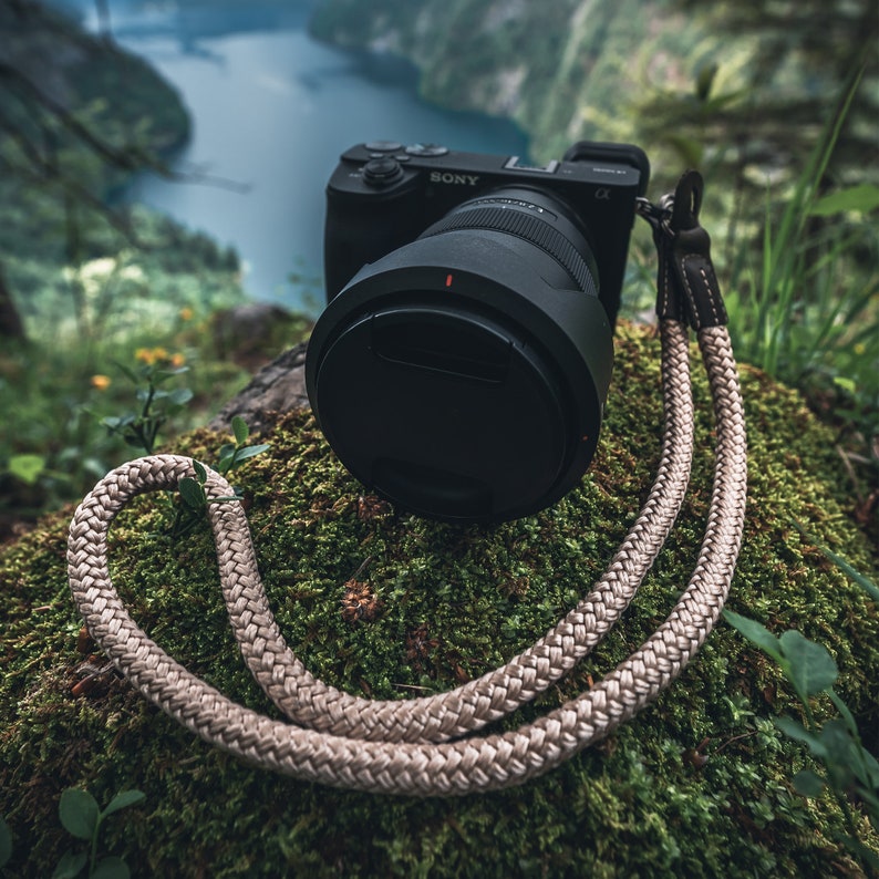 May include: A black camera with a lens and a beige camera strap. The camera is sitting on a bed of green moss.