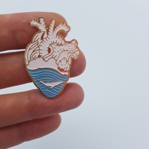 May include: A heart-shaped enamel pin with a gold border. The top half is white and pink, the bottom half is blue with a white whale. The pin is held between two fingers against a white background.