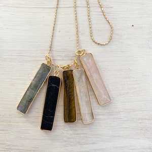 May include: A gold chain necklace with five different colored gemstone bar pendants. The pendants are in shades of grey, black, brown, white, and pink.