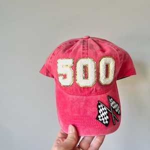 May include: A pink baseball cap with a white "500" embroidered on the front in gold lettering. The cap also has two black and white checkered flags on the side.