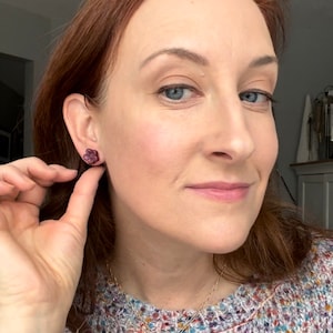 May include: A close-up of a person wearing a pair of dark red flower-shaped stud earrings. The earrings are being touched by a hand. A gold necklace with a small pendant is also visible.