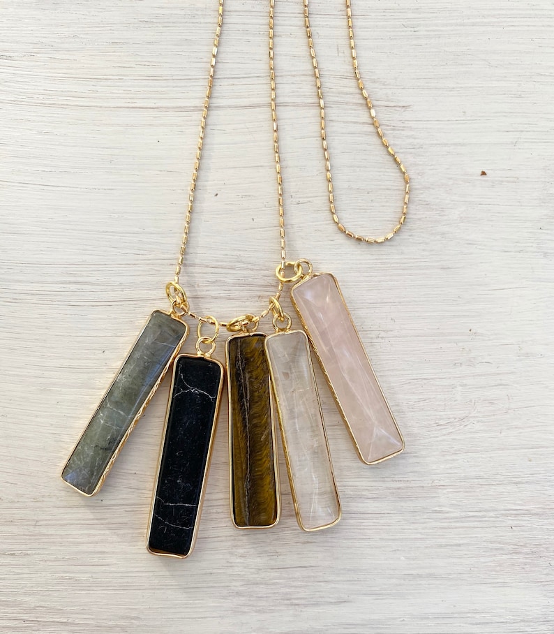 May include: A gold chain necklace with five different colored gemstone bar pendants. The pendants are in shades of grey, black, brown, white, and pink.