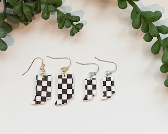 May include: Four pairs of earrings with a black and white checkered pattern in the shape of the state of Indiana. The earrings have gold and silver colored hooks. The earrings are displayed on a white surface with green foliage.