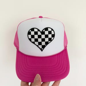 May include: A pink trucker hat with a white front panel featuring a black and white checkered heart design. The hat has a pink brim and mesh sides, and a pink button on top.
