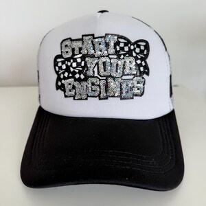 May include: A white trucker hat with a black brim. The hat features the text "START YOUR ENGINES" in silver sequins, with a checkered flag design. The hat is designed for motorsports enthusiasts.