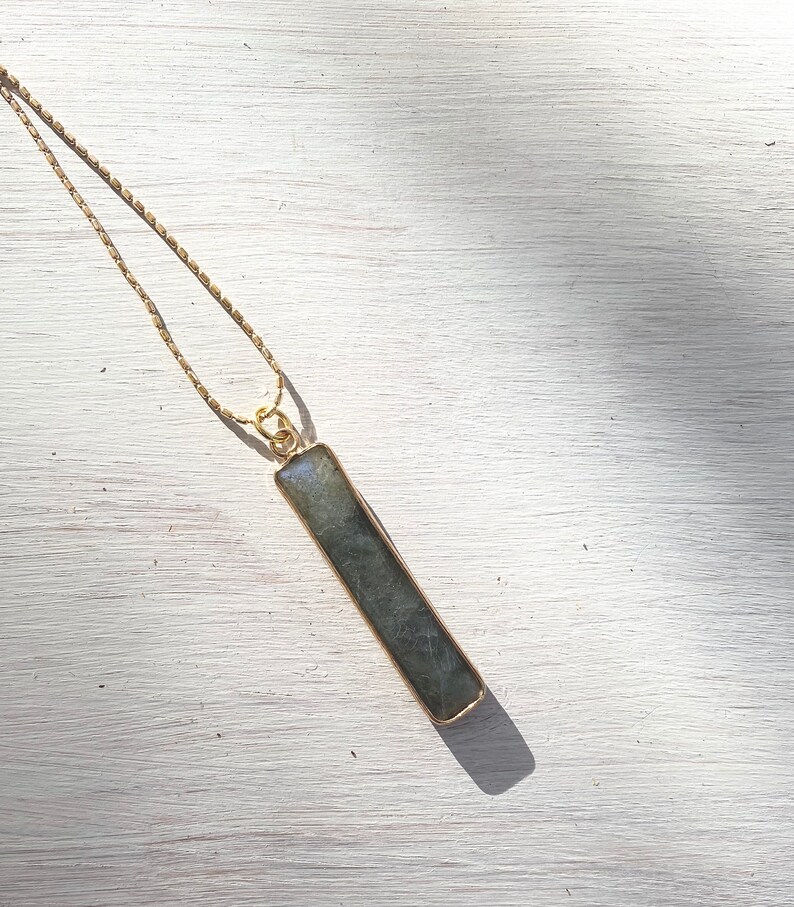 May include: A gold chain necklace with a long, rectangular, dark grey labradorite pendant.