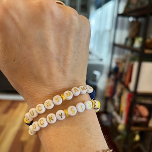 May include: Two white beaded bracelets with gold letters spelling "LUCKY MAMA" and "LUCKY SIS".