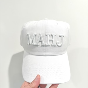 May include: A white baseball cap with the word "MAHJ" embroidered on the front in raised, white letters. The cap has a curved brim and a structured crown. The background is a plain white wall.