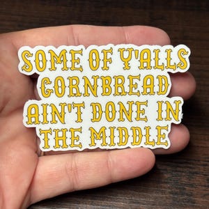 May include: A white sticker with yellow text that reads "SOME OF Y'ALLS CORNBREAD AIN'T DONE IN THE MIDDLE." The text has a decorative font and is outlined in yellow. The sticker is held in a hand.
