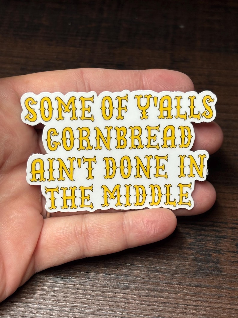 May include: A white sticker with yellow text that reads "SOME OF Y'ALLS CORNBREAD AIN'T DONE IN THE MIDDLE." The text has a decorative font and is outlined in yellow. The sticker is held in a hand.