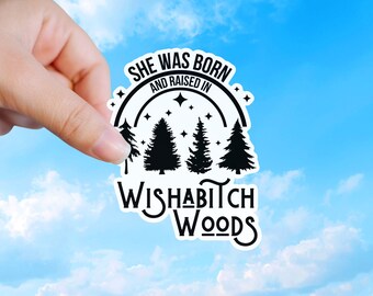 May include: A white die-cut sticker featuring black text that reads SHE WAS BORN AND RAISED IN WISHABITCH WOODS. The design includes a black arched line, four black pine tree silhouettes, and small four-pointed star icons against a white background.