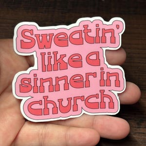 May include: A pink sticker with the text "Sweatin' like a sinner in church" in red block letters. The sticker has a white border. The sticker is held in a person's hand.