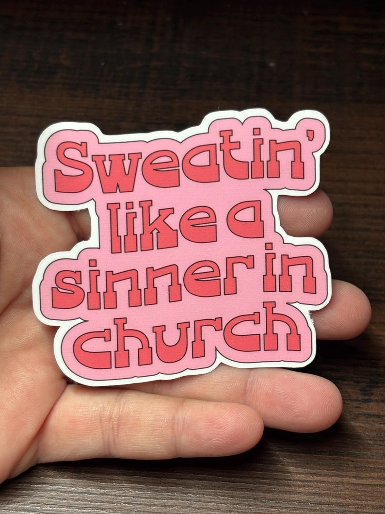 May include: A pink sticker with the text "Sweatin' like a sinner in church" in red block letters. The sticker has a white border. The sticker is held in a person's hand.