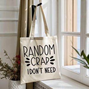 May include: Cream-colored tote bag with the text "RANDOM CRAP I DON'T NEED" in black. The bag has long, beige handles and is displayed near a window with a neutral-toned curtain and a potted plant.