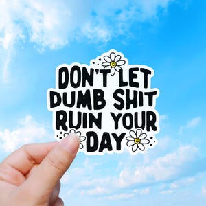 May include: A white sticker with black text that reads "DON'T LET DUMB SHIT RUIN YOUR DAY." The sticker has daisy accents and is held against a blue sky background.