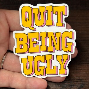 May include: A sticker with the text "QUIT BEING UGLY" in yellow bubble letters with a pink outline. The sticker is on a white background. The sticker is held in a hand.