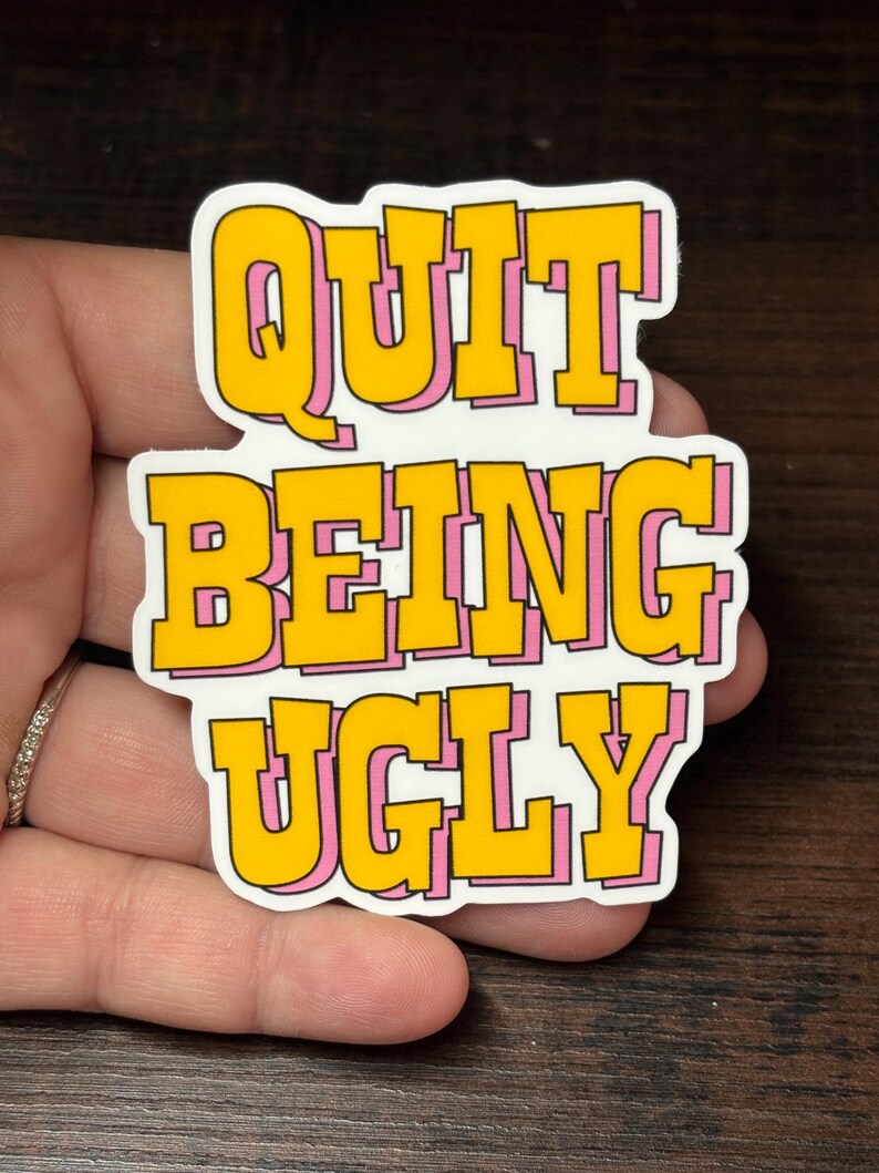 May include: A sticker with the text "QUIT BEING UGLY" in yellow bubble letters with a pink outline. The sticker is on a white background. The sticker is held in a hand.