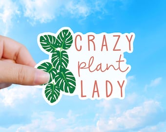 May include: A die-cut sticker featuring a cluster of green monstera leaves on the left side and the text CRAZY plant LADY in a reddish-brown font on the right side, all set against a white background with a thin white border outline.