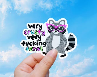 Puede incluir: Una pegatina de vinilo con un mapache gris y blanco que lleva gafas de color rosa brillante. El texto dice very crusty very fucking feral en letras negras, moradas, azules y verdes. La pegatina tiene un borde blanco y forma troquelada.