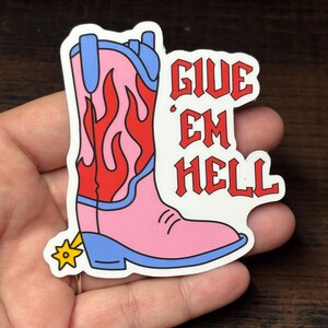 May include: A sticker depicting a pink cowboy boot with red flames and blue detailing. The boot has a gold spur and the red text "GIVE EM HELL". The sticker is held in a hand.