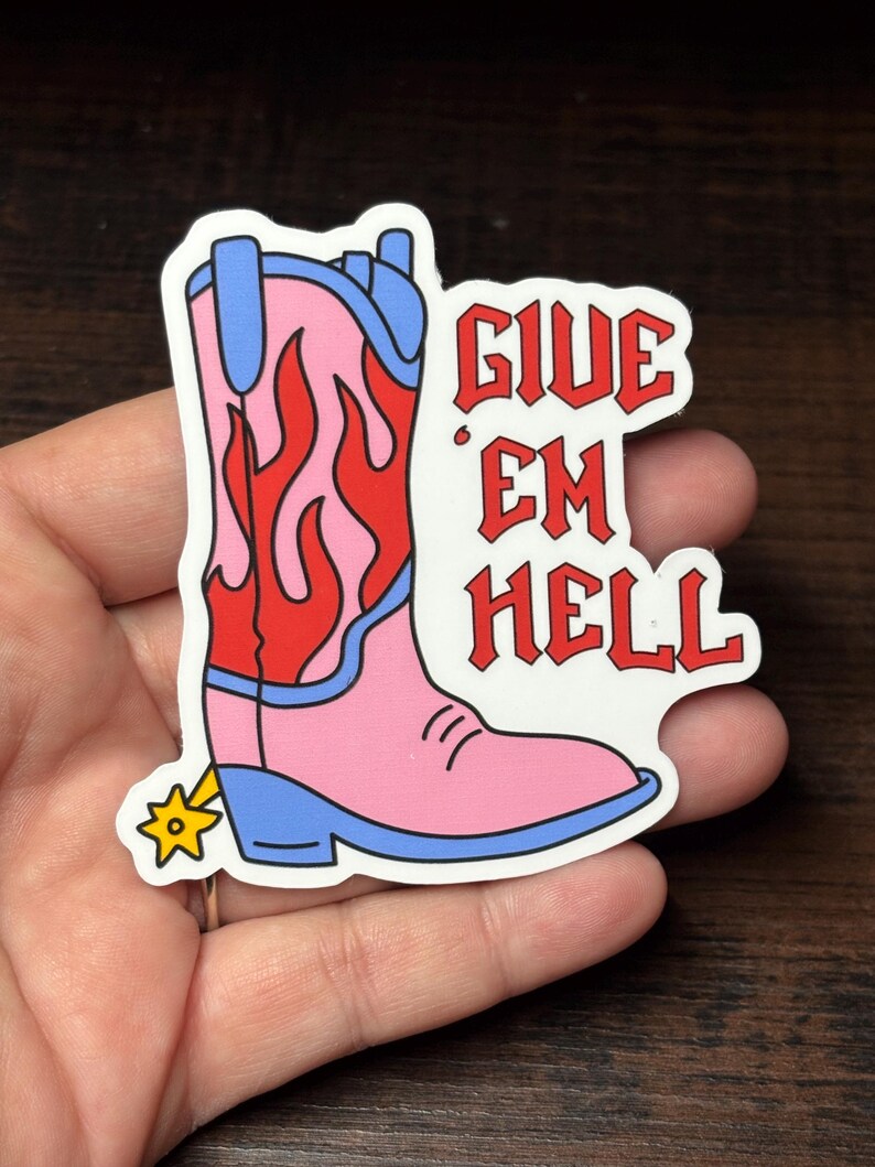 May include: A sticker depicting a pink cowboy boot with red flames and blue detailing. The boot has a gold spur and the red text "GIVE EM HELL". The sticker is held in a hand.