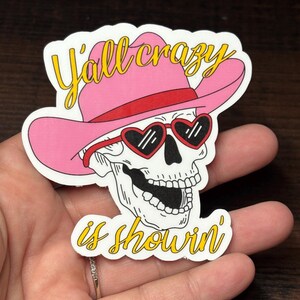 May include: A sticker featuring a skull wearing a pink cowboy hat and heart-shaped sunglasses. The text "Y'all crazy is showin'" is written in yellow script. The design is outlined in black and white.