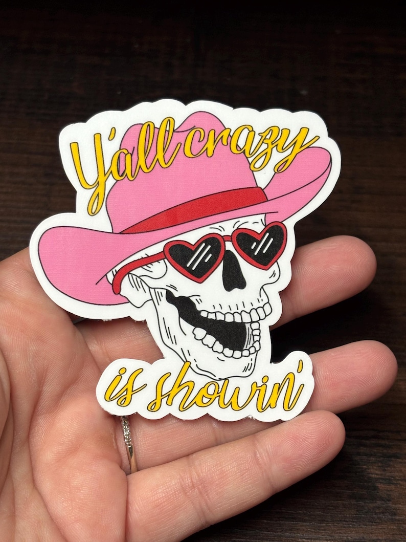 May include: A sticker featuring a skull wearing a pink cowboy hat and heart-shaped sunglasses. The text "Y'all crazy is showin'" is written in yellow script. The design is outlined in black and white.