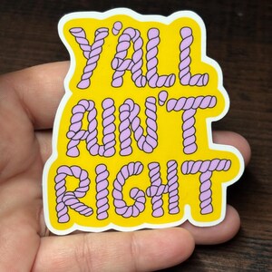 May include: A yellow sticker with the words "Y'ALL AIN'T RIGHT" in purple rope-style lettering. The sticker has a white border and is held in a hand against a dark wood background.