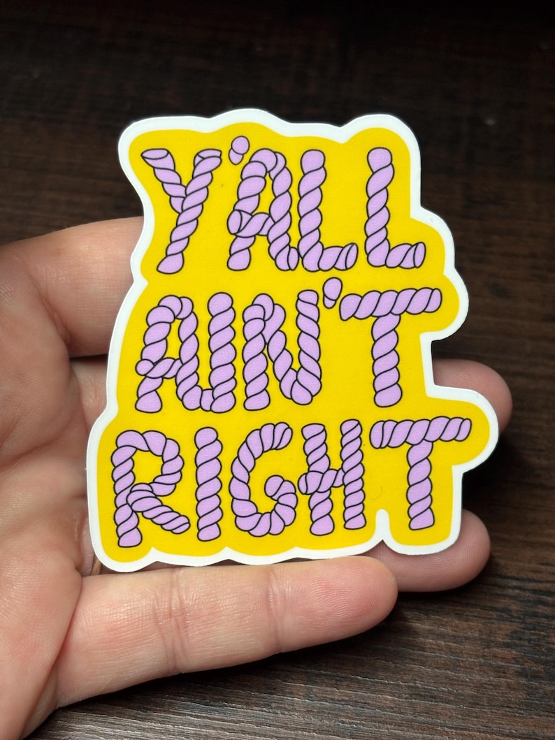 May include: A yellow sticker with the words "Y'ALL AIN'T RIGHT" in purple rope-style lettering. The sticker has a white border and is held in a hand against a dark wood background.