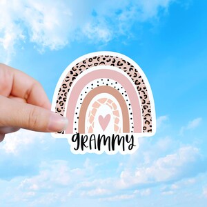 May include: A die-cut sticker featuring a rainbow design with four arches in shades of pink, tan, and brown. The outer arch has a leopard print, the inner arches have dots and a giraffe pattern, and a pink heart sits in the center with the word GRAMMY.