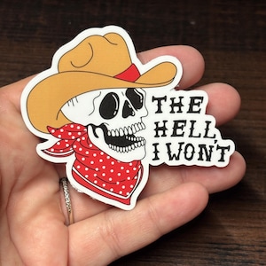 May include: A sticker depicting a skull wearing a cowboy hat and a red polka dot bandana. The text "THE HELL I WON'T" is printed in black. The sticker is held in a hand.