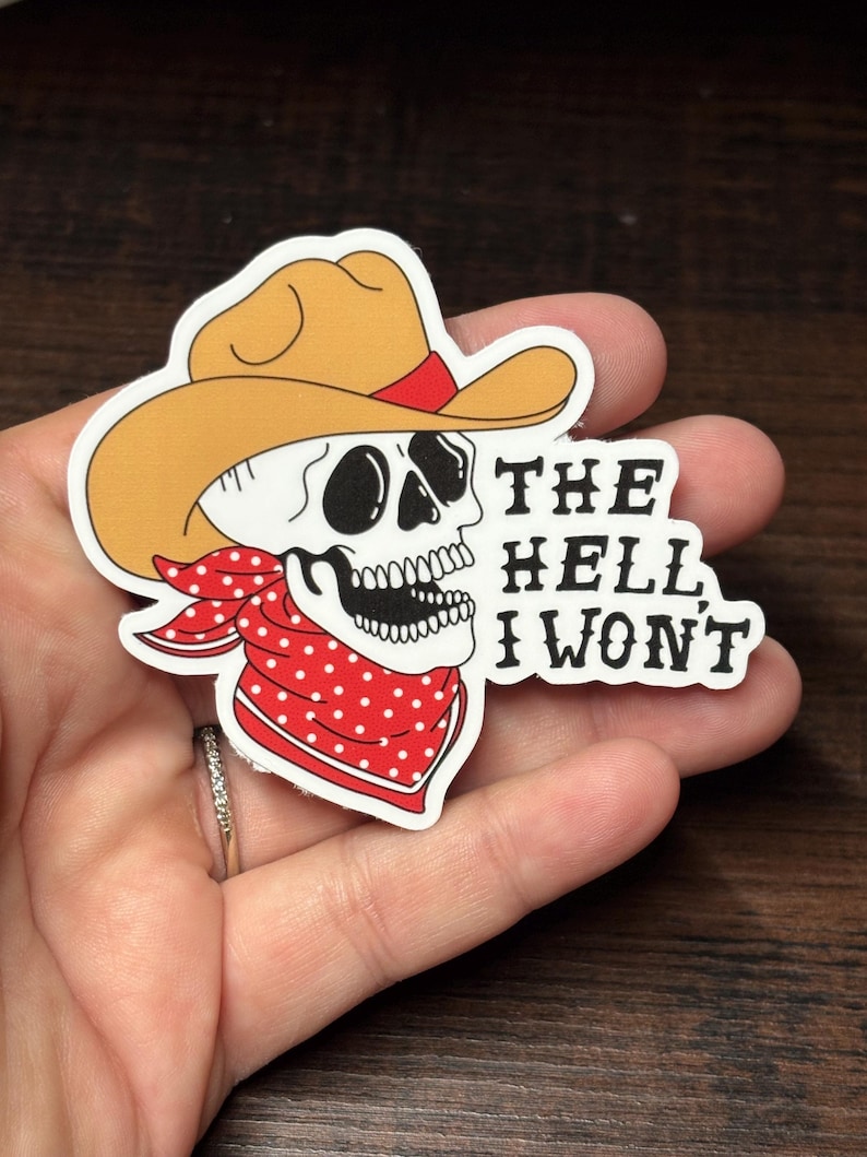 May include: A sticker depicting a skull wearing a cowboy hat and a red polka dot bandana. The text "THE HELL I WON'T" is printed in black. The sticker is held in a hand.