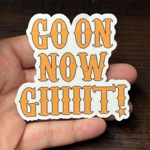 May include: A white sticker with orange text that reads "GO ON NOW GIIIIIT!" in a bold, vintage font. A small star is next to the exclamation mark. The sticker is held in a hand.