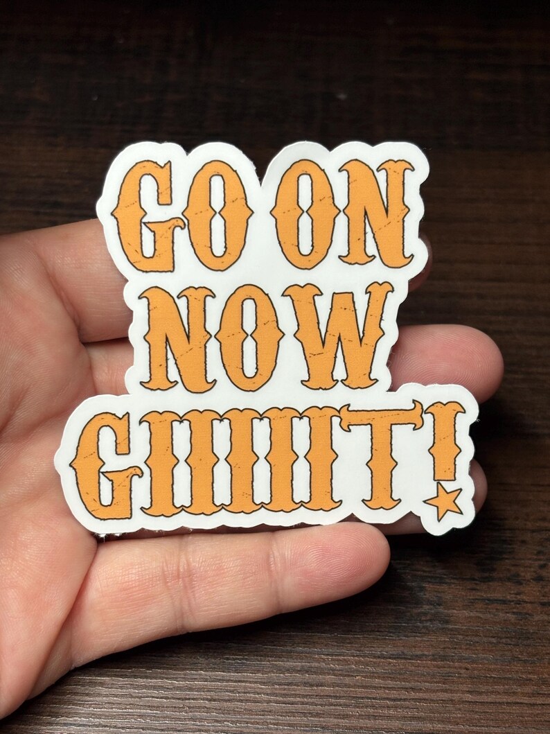 May include: A white sticker with orange text that reads "GO ON NOW GIIIIIT!" in a bold, vintage font. A small star is next to the exclamation mark. The sticker is held in a hand.