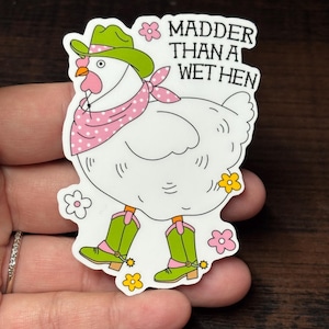 May include: A white chicken sticker wearing a green cowboy hat, pink polka dot bandana, and green cowboy boots. The sticker has the text "Madder Than A Wet Hen" in white. The sticker is decorated with pink and yellow flowers.