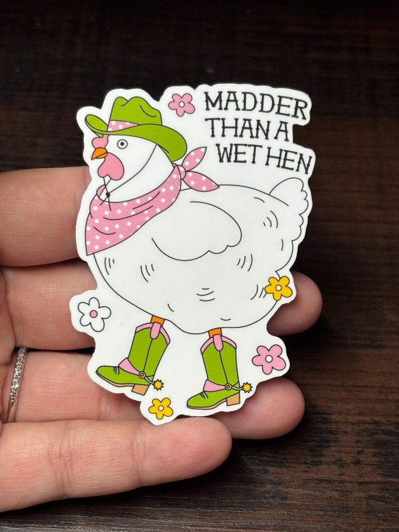 May include: A white chicken sticker wearing a green cowboy hat, pink polka dot bandana, and green cowboy boots. The sticker has the text "Madder Than A Wet Hen" in white. The sticker is decorated with pink and yellow flowers.