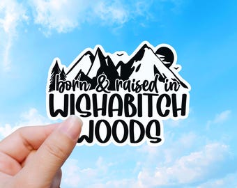 May include: A die-cut vinyl sticker featuring a black and white graphic of mountain peaks, pine trees, and a sun. The text reads "born & raised in WISHABITCH WOODS" in a combination of cursive and bold, sans-serif black lettering on a white background.