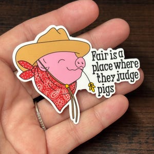 May include: A sticker depicting a cartoon pig in a cowboy hat and red bandana. The pig holds a piece of straw. The text on the sticker says "Fair is a place where they judge pigs."