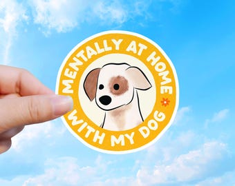 May include: A circular sticker featuring a white dog with a brown ear and eye patch in the center. The background is yellow with the white text MENTALLY AT HOME WITH MY DOG written in a circular arc, accented by a small orange flower icon.