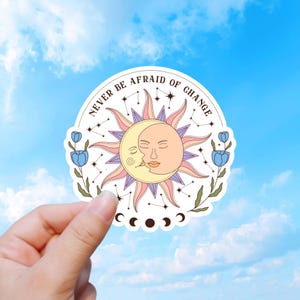 May include: A circular sticker featuring a sun and moon face illustration with pink and yellow rays. The text NEVER BE AFRAID OF CHANGE is printed in an arc above. Blue tulip flowers on green stems flank the sides, with small star constellations.