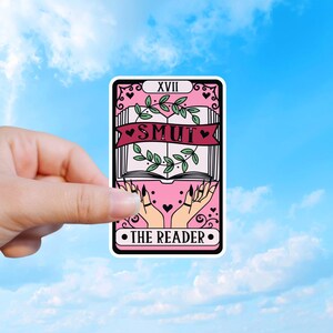Könnte beinhalten: Ein rechteckiger Aufkleber im Design einer Tarotkarte mit rosa Hintergrund. Er zeigt ein offenes Buch mit grünen Blättern, ein dunkelrosa Banner mit dem Text SMUT und zwei Hände darunter. Oben steht XVII und unten steht THE READER.