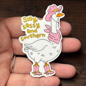 May include: A white goose sticker wearing a pink cowboy hat, bandana, and boots. The sticker features the text "Silly, sassy, and southern" in yellow. The goose has an orange beak and is wearing pink boots.