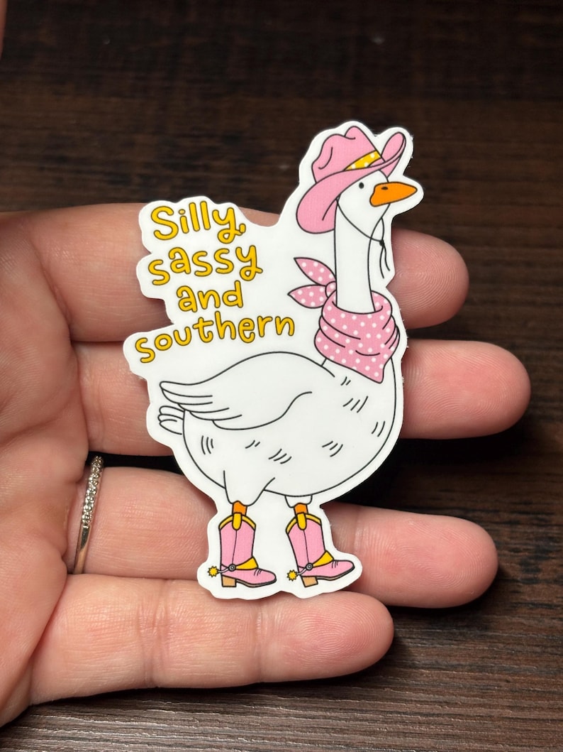 May include: A white goose sticker wearing a pink cowboy hat, bandana, and boots. The sticker features the text "Silly, sassy, and southern" in yellow. The goose has an orange beak and is wearing pink boots.